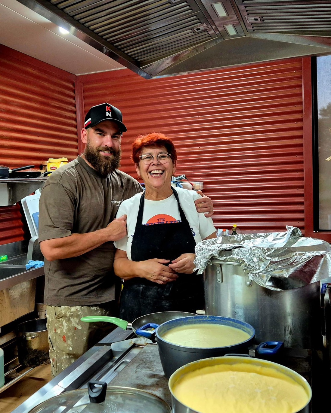 Aunty Donna smiling in the kitchen beside family and large pots of food.